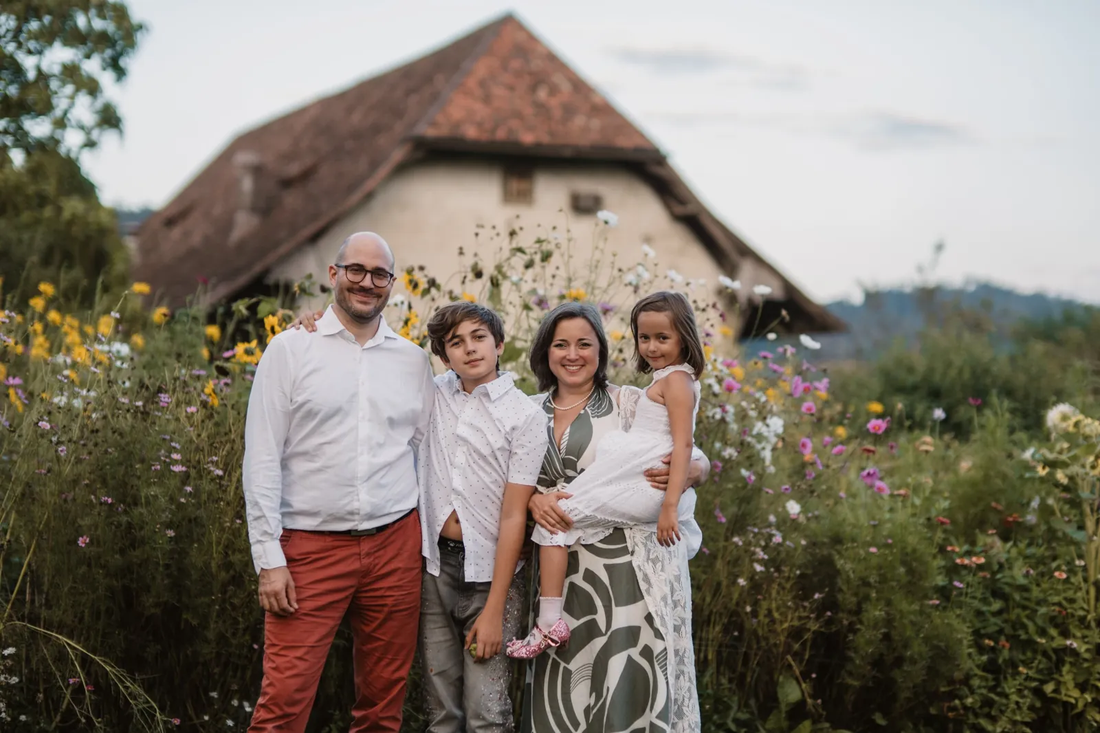 Familie mit zwei Kindern steht lachend vor einem alten Bauernhaus zwischen hohen Sommerblumen
