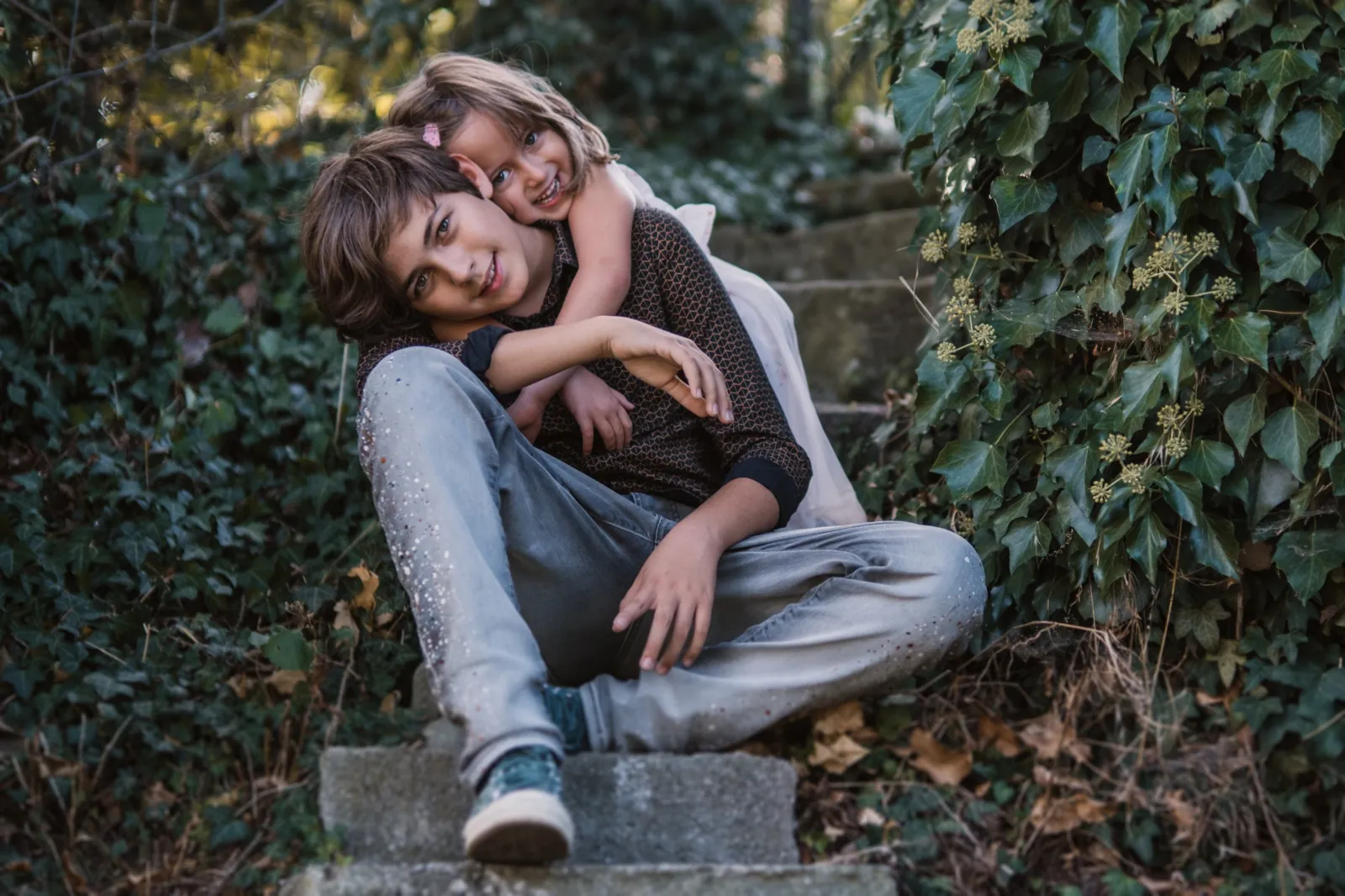 Die kleine Schwester umarmt ihren Bruder von hinten, beide sitzen lachend auf einer Steintreppe im Efeu