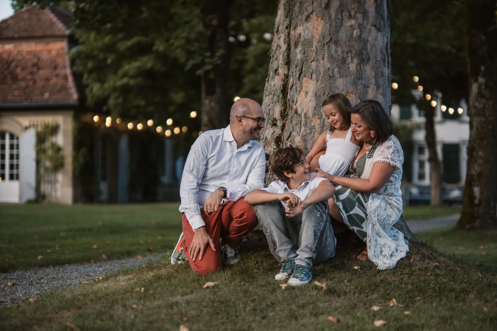 Die Familie sitzt lachend am Fuss eines grossen Baumes und schaut sich gegenseitig an